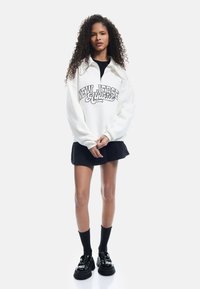 Felpa bianca con grafica "NEW JERSEY Authentic," abbinata a una gonna nera, calze fino al ginocchio nere e scarpe nere lucide.
