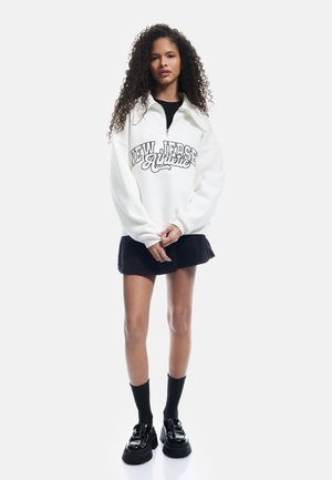 Sudadera blanca con el gráfico "NEW JERSEY Authentic," combinada con una falda negra, medias hasta la rodilla negras y zapatos negros brillantes.