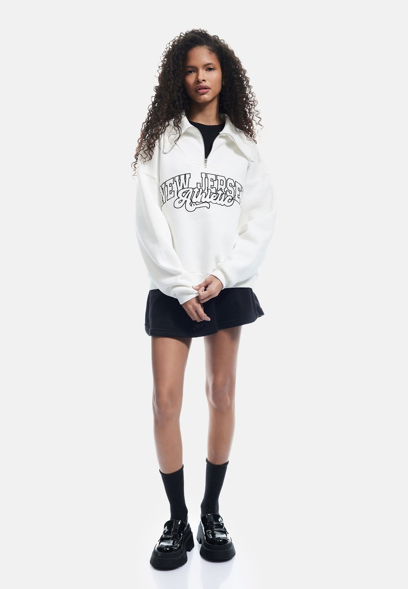 Felpa bianca con grafica "NEW JERSEY Authentic," abbinata a una gonna nera, calze fino al ginocchio nere e scarpe nere lucide.