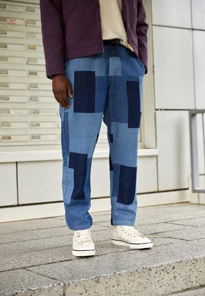 Personne portant un pantalon en jean patchwork bleu, des baskets blanches, un haut beige et une veste violette, debout sur des marches en pierre à l'extérieur.