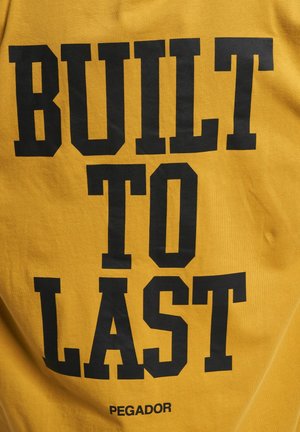 Gul skjorte i bomull med fet svart tekst som sier "BUILT TO LAST" med "PEGADOR"-logoen under. Myk tekstur, uformell design.