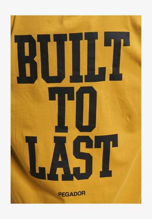 Gul skjorte i bomull med fet svart tekst som sier "BUILT TO LAST" med "PEGADOR"-logoen under. Myk tekstur, uformell design.