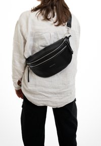 Personne portant une chemise blanche à manches longues et un pantalon noir avec un sac en cuir noir bandoulière, orné de fermetures éclair argentées et du texte « EXPATRIÉ ».