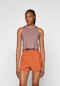 Donna con la pelle scura e la coda di cavallo che indossa un top corto senza maniche color malva e pantaloni sportivi arancioni, in piedi contro uno sfondo chiaro e uniforme.