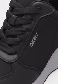 Μαύρο αθλητικό παπούτσι με χοντρά κορδόνια, το λογότυπο DKNY στο πλάι, λείες και υφασμένες επιφάνειες, και λευκή σόλα ορατή στο κάτω δεξιά μέρος.