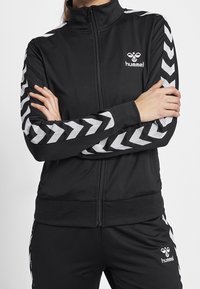 Black zip-up jacket made of smooth fabric, featuring white chevron stripes on sleeves and a small logo on the chest.