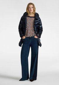 Giacca a vento blu navy con cappuccio, maglione a fantasia con cerchi in tonalità di bordeaux e crema, abbinato a jeans di denim scuri e larghi.