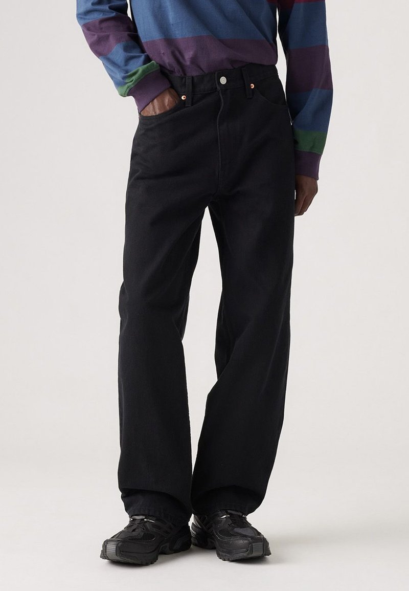 Jeans neri a gamba larga in denim, con vita alta, tasche frontali e cuciture a contrasto. Abbinati a una camicia a maniche lunghe multicolore.
