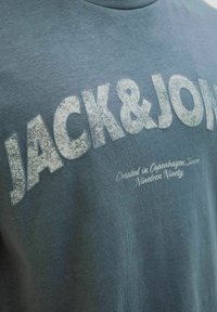 Bomulds t-shirt i dyb teal med stor hvid tekst "JACK&JON" og mindre tekst "Skabt i København Siden nitten-nitten." Glat tekstur.