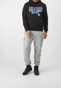 Hoodie noir avec le texte "Los Angeles Dodgers" en bleu et rouge, associé à un pantalon de survêtement gris avec des zippers sur les côtés et des baskets noires.