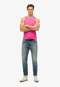 Top sin mangas rosa, jeans de mezclilla azules, zapatillas blancas y beige, postura de pie con los brazos cruzados, accesorios mínimos, fondo liso.