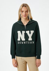 Sudadera verde con cuello alto, con cierre de cremallera y texto blanco "NY DOWNTOWN". Los puños y el dobladillo acanalados añaden textura.