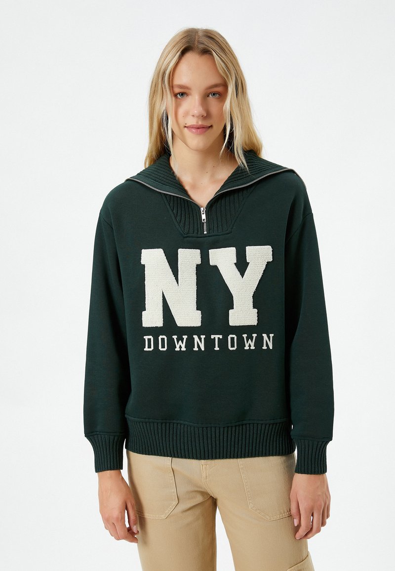 Sudadera verde con cuello alto, con cierre de cremallera y texto blanco "NY DOWNTOWN". Los puños y el dobladillo acanalados añaden textura.