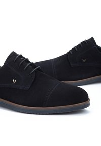 Un paio di scarpe eleganti in pelle scamosciata nera con lacci, suola marrone e una sottile cucitura decorativa su uno sfondo bianco.