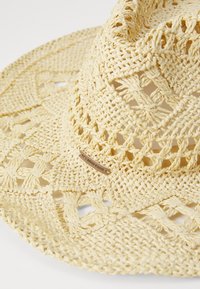 Sombrero de paja tejido en amarillo claro, con un diseño de tejido abierto, ala ancha y una etiqueta de tela marrón en el costado.