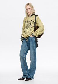 Olīvu zaļš kapuces krekls ar grafisku drukājumu, virsū uzsliets uz režģa krekla, apvienots ar gaiši ziliem taisnām kājām džinsiem un smailām melnām kurpēm.