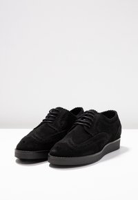 Scarpe in suede nero con design brogue, presentano una punta arrotondata, cuciture intricate e una suola a piatto bassa. Chiusura con lacci standard.