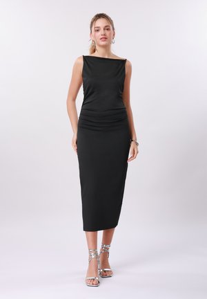 Γυναίκα όρθια, φοράει μαύρο μίντι φόρεμα χωρίς μανίκια με λαιμόκοψη καραβάκι και ασημένια ψηλοτάκουνα σανδάλια με λουράκια σε φωτεινό φόντο.