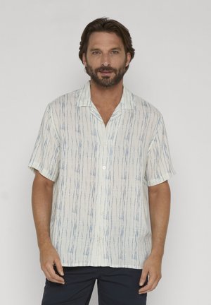 Homme aux cheveux bruns courts et à la barbe, portant une chemise blanche à manches courtes à boutons avec des rayures verticales bleues et un pantalon foncé.
