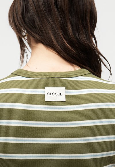 Άτομο που φαίνεται από πίσω φοράει πουκάμισο με οριζόντιες ρίγες σε ελιά και λευκό, με ραμμένη ορθογώνια ετικέτα που γράφει "CLOSED" στη πάνω πίσω πλευρά του γιακά.