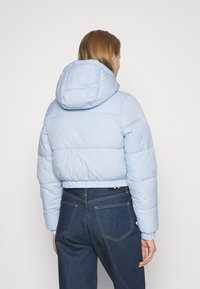 Žena v světle modré péřové bundě s kapucí a tmavě modrých vysokých džínách, stojící zády k objektivu na jednoduchém bílém pozadí.