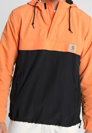 Mężczyzna noszący dwukolorową pomarańczowo-czarną kurtkę Carhartt z kapturem, z przednimi kieszeniami na suwak i sznurkami.