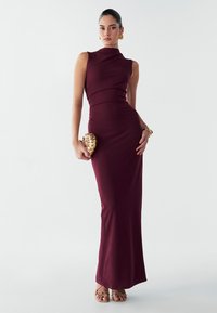 Vīna sarkana bez piedurknēm pieguloša kleita ar augstu kakla izgriezumu, gludu tekstūru un pieguļošu siluetu. Rotāta ar zelta čaumalas formas somiņu.