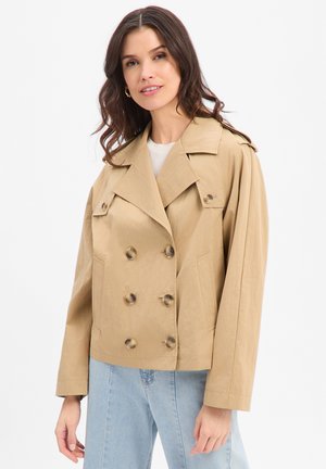 Frau mit welligem braunem Haar, die goldene Creolen trägt, einen beigen, doppelreihig geknöpften, kurzen Trenchcoat, ein weißes Oberteil und hellblaue Jeans, lächelnd.