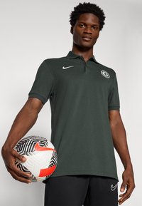 Mørkegrøn polo skjorte med krave, der har et hvidt Nike-logo og et Chelsea FC-mærke. Han holder en mønstret sort-hvid fodbold.