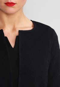 Texturerad svart jacka med rund krage, något öppen fram, och en sluten vävstruktur. Bärs över en v-ringad topp.