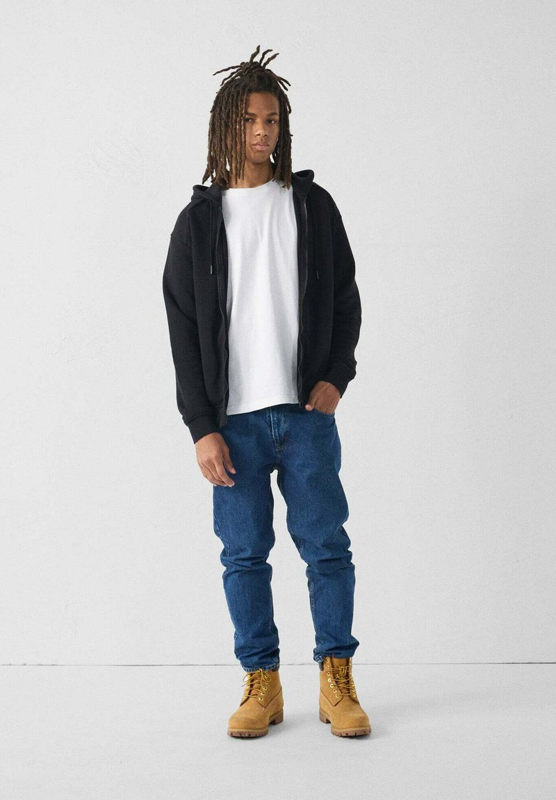 Sudadera negra con cremallera, camiseta blanca, jeans azules y botas de trabajo amarillas. El modelo está de pie con las manos en los bolsillos sobre un fondo liso.