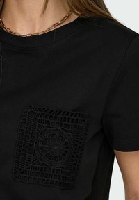 Svart T-shirt med en texturerad, svart spetsficka med ett cirkulärt mönster. Klassisk rund halsringning och korta ärmar.