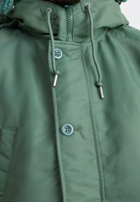 Grüne Kapuzenjacke mit glatter Textur, ausgestattet mit Reißverschluss, Knopfdetails und verstellbaren Kordeln an der Kapuze.