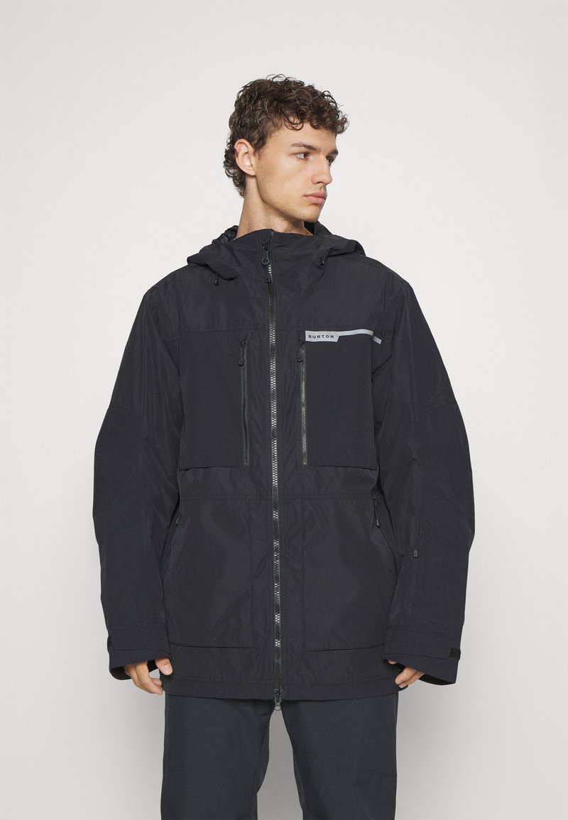 Veste noire imperméable avec fermeture à glissière, capuche réglable et plusieurs poches zippées. Présente un logo gris et une coupe décontractée.
