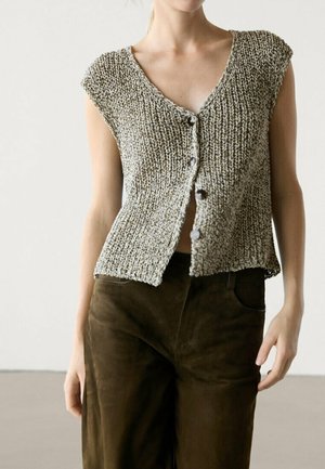 Femme portant un gilet gris en maille sans manches, boutonné, et un pantalon ample marron foncé, debout devant un fond neutre.