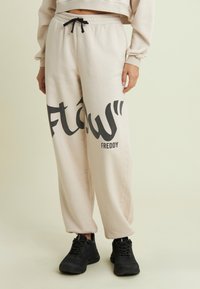 Pantalones de chándal color crema con una impresión gráfica negra que dice "flow" y "Freddy" en el costado; pretina elástica con un cordón negro.