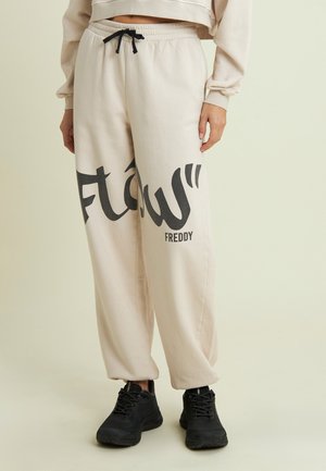 Pantalons de survêtement couleur crème avec un imprimé graphique noir portant les mots "flow" et "Freddy" sur le côté ; taille élastique avec un cordon de serrage noir.