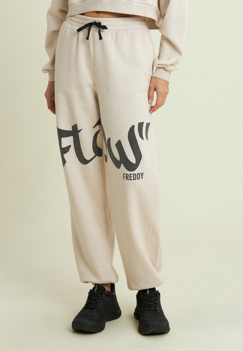 Pantalones de chándal color crema con una impresión gráfica negra que dice "flow" y "Freddy" en el costado; pretina elástica con un cordón negro.