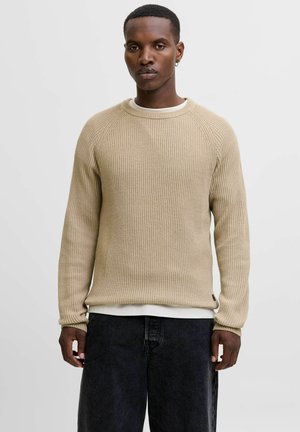 Beige rib gebreide trui met een ronde hals en raglanmouwen, bovenop een wit T-shirt, gecombineerd met donkere spijkerbroek.