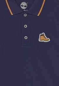 Námořnická modrá polo košile s oranžovými akcenty na límci, tříknoflíkovou légou a žlutým a černým vyšitým logem boty na levé hrudi.