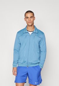 Chaqueta de color azul claro con cierre de cremallera y cuello alto, fabricada en tejido suave, que presenta rayas de contraste y dos bolsillos laterales.