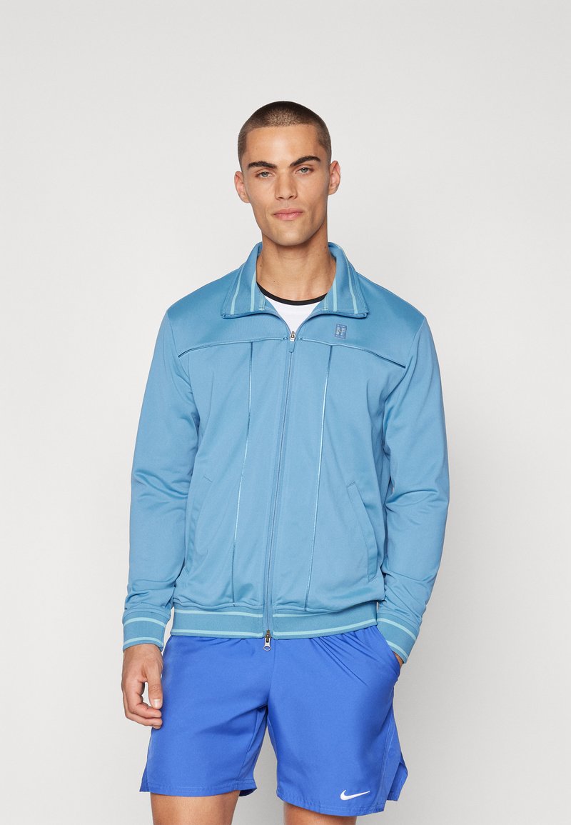 Chaqueta de color azul claro con cierre de cremallera y cuello alto, fabricada en tejido suave, que presenta rayas de contraste y dos bolsillos laterales.