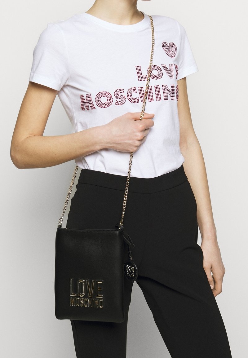 Donna che indossa una maglietta bianca "Love Moschino" con borchie e pantaloni neri, mentre tiene una borsa a tracolla nera "Love Moschino" con catena dorata.