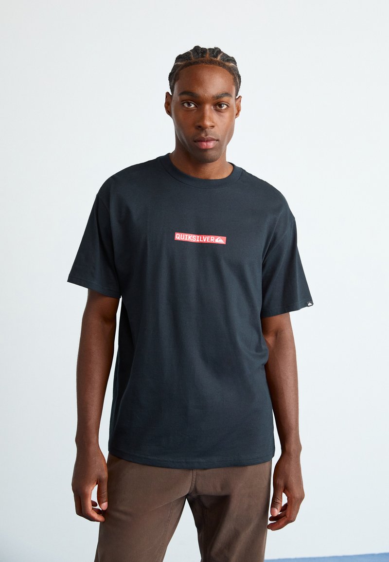 Jeune homme avec des tresses portant un t-shirt noir Quiksilver et un pantalon marron, se tenant devant un fond clair uni.