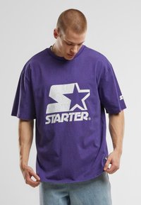 T-shirt en coton violet avec un grand logo blanc "STARTER" sur le devant, des manches courtes et une coupe décontractée.