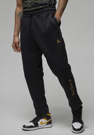 Pantalon de survêtement noir avec logo Jumpman jaune et texte "PARIS", porté avec des baskets Nike noires, blanches et jaunes. Tatouages visibles sur les bras.