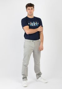 T-shirt in cotone navy con grafica nautica, abbinato a pantaloni in cotone grigio chiaro. Il modello è in piedi con le braccia incrociate, indossando sneakers bianche.