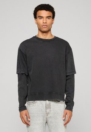 Junger Mann mit lockigem Haar, der ein schwarzes Langarm- und Kurzarmshirt in Lagen trägt, kombiniert mit hellgrauen Jeans, steht und schaut nach vorne.