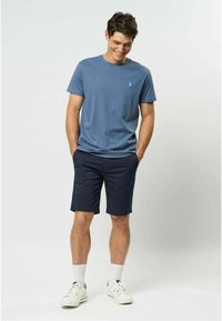 Niebieska bawełniana koszulka z małym zielonym logo, zestawiona z granatowymi szortami i białymi trampkami. Model stoi z rękami w kieszeniach, uśmiechając się.