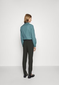 Türkisfarbenes Hemd mit langen Ärmeln, kombiniert mit dunkelgrünen Hosen. Hat einen taillierten Schnitt und eine glatte Textur. Schwarze Slipper runden das Outfit ab.